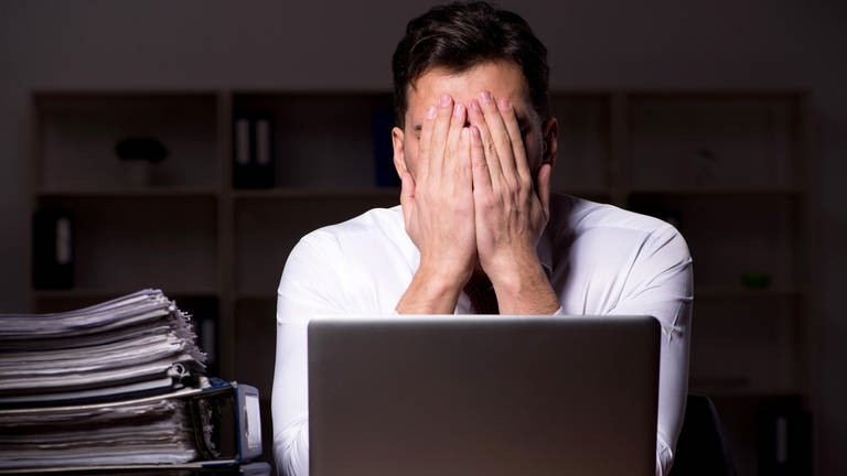 Arbeitssucht: Ein Mann sitzt nachts an seinem Schreibtisch vor einem Notebook und einem Stapel Aktenordnern | Arbeitssucht, Arbeit und Gesundheit