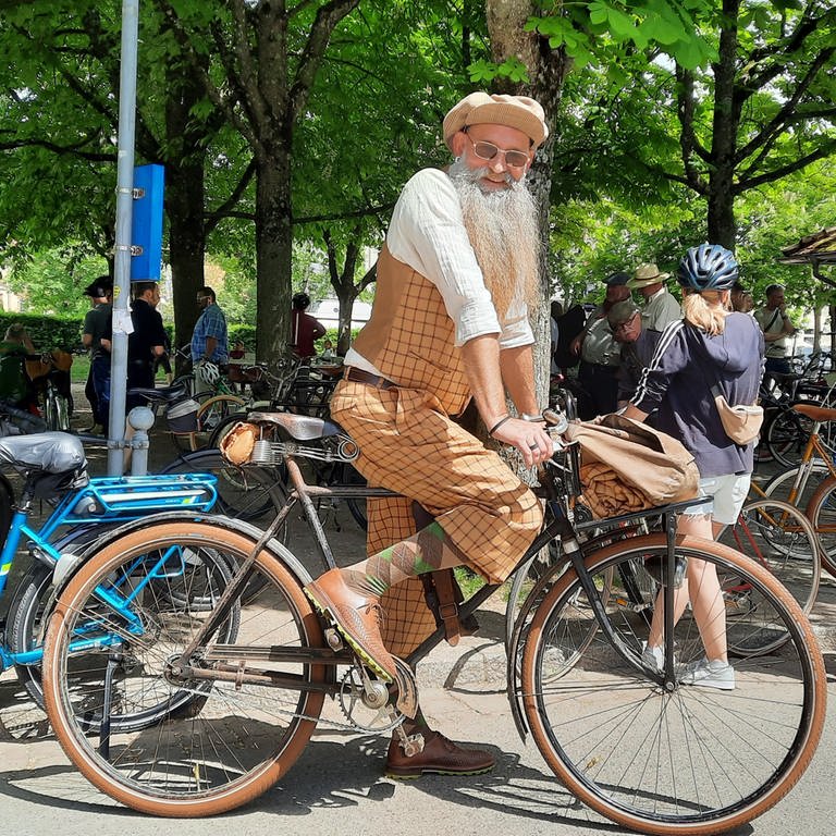 Ein Mann mit langem weißem Bart in karierter Kleidung auf einem Retro-Bike lacht in die Kamera.