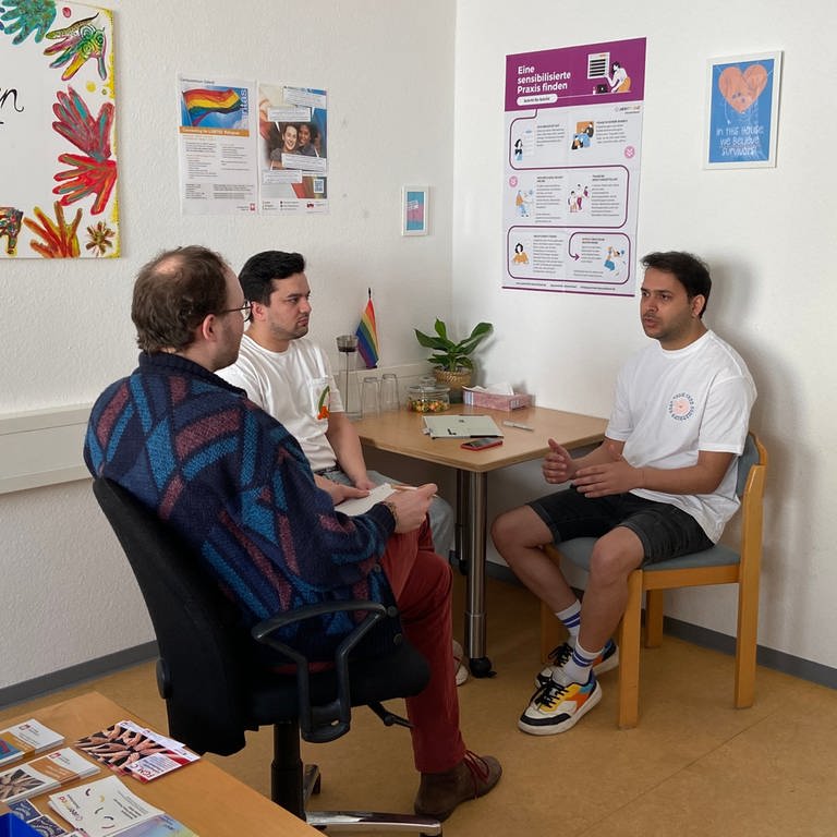 Zwei queere Männer sitzen bei Konsti Hahn in der Beratungsstelle für Flüchtlinge der Caritas in Mainz.
